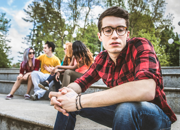 Das Bild zeigt einen jungen Mann mit Brille und kariertem Hemd, der auf einer Treppe sitzt und nachdenklich in die Kamera blickt. 